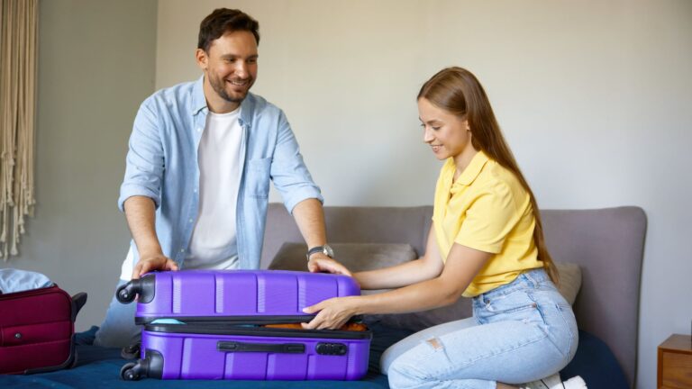 Ein Mann und eine Frau lächeln, während sie einen großen lila Koffer auf dem Bett packen und dabei Gegenstände von der perfekten Packliste abhaken, während sie sich auf ihre Reise vorbereiten.